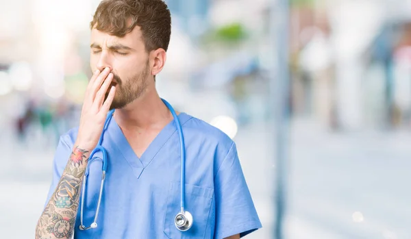 Young handsome nurse man wearing surgeon uniform over isolated background bored yawning tired covering mouth with hand. Restless and sleepiness.