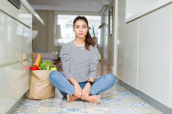 Young woman sitting on the kitchen floor with a paper bag full of fresh groceries with serious expression on face. Simple and natural looking at the camera.