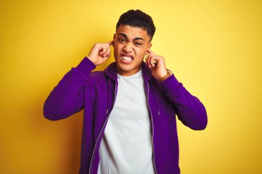 Genç Brezilyalı adam mor sweatshirt yüksek sesle gürültü için rahatsız ifade ile parmakları ile kulakları kapsayan izole sarı arka plan üzerinde duran giyiyor. Sağır kavramı.