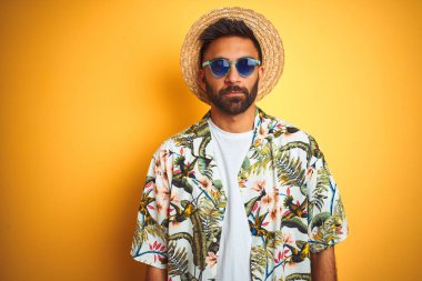 Tatilde Hintli adam yüzünde ciddi ifade ile izole sarı arka plan üzerinde çiçek gömlek şapka güneş gözlüğü giyiyor. Kameraya basit ve doğal bir bakış.