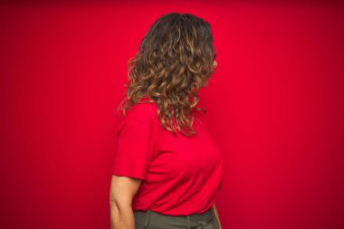 Kırmızı izole arka plan üzerinde kıvırcık saçlı orta yaş yaşlı kadın yan bakıyor, kendine güvenen gülümseme ile doğal yüz ile profil poz dinlenmek.