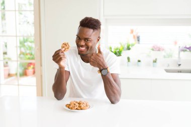 Genç afro-amerikan adam büyük bir gülümseme ok işareti yapıyor mutlu çikolata parçacıkları çerezleri yeme, parmakları ile başparmak, mükemmel işareti