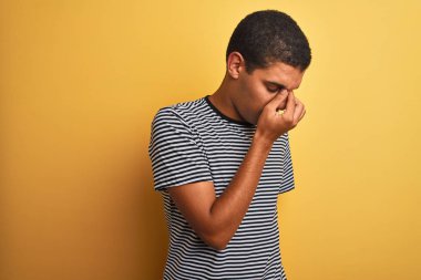 Genç yakışıklı Arap adam izole sarı arka plan yorgun sürtünme burun ve gözler üzerinde lacivert çizgili t-shirt giyen yorgunluk ve baş ağrısı hissi. Stres ve hayal kırıklığı kavramı.
