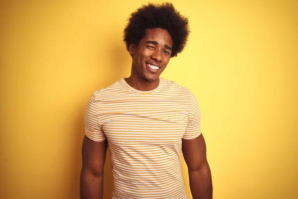American man with afro hair wearing striped t-shirt standing over isolated yellow background winking looking at the camera with sexy expression, cheerful and happy face.