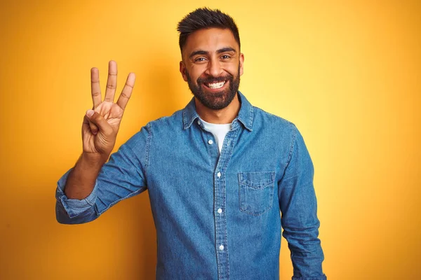 孤立した黄色の背景の上に立つデニムシャツを着た若いインド人男性は 自信を持って幸せに微笑みながら 3番の指で見せ 指を上げている — ストック写真