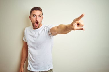 Genç yakışıklı adam izole arka plan üzerinde rahat beyaz t-shirt giyen parmak ile işaret önünde şaşırttı, açık ağız şaşkın ifade, ön bir şey