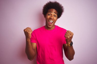 Afro saçlı, tişört giyen genç amerikalı adam izole edilmiş pembe arka plan üzerinde durarak kollarını kaldırıp gözleri açık bir şekilde başarıya uğruyor. Kazanan kavramı.