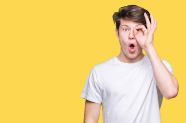 Sürpriz yüz, göz parmaklarının arasından seyir ile ok jest yapıyor izole arka plan üzerinde rahat beyaz t-shirt giyen genç yakışıklı adam şok. İmansız ifade.
