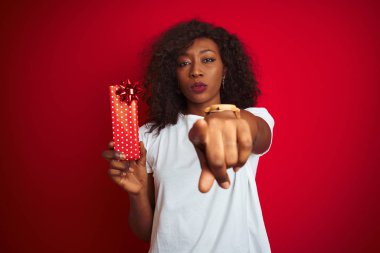 Genç afro-amerikan kadın kamera ve size parmak ile işaret izole kırmızı arka plan üzerinde duran sevgililer hediye tutan, el işareti, önden olumlu ve kendinden emin jest