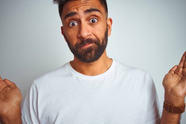 Genç hintli adam izole beyaz arka plan üzerinde duran t-shirt giyen habersiz ve kolları ve elleri kaldırdı ile karışık ifade. Şüphe kavramı.