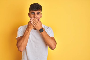 İzole edilmiş sarı arka planın üzerinde duran beyaz tişört giyen hintli genç adam hata için elleri ile ağız örtüyor şok etti. Gizli kavram.