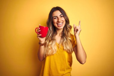 Genç güzel kadın sarı izole arka plan üzerinde kahve kırmızı fincan tutan çok mutlu yan el ve parmak ile işaret