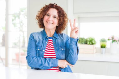 Evde kot ceket giyen kıvırcık saçlı orta yaş yaşlı kadın parmaklarıyla zafer işareti yaparak kameraya göz kırparak mutlu yüz ile gülümsüyor. İki numara.