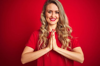 Kırmızı izole arka plan üzerinde duran temel t-shirt giyen genç güzel kadın birlikte emin gülümseyerek af isteyen elleri ile dua.