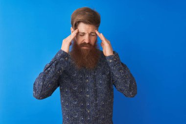 Genç kızıl saçlı İrlandalı adam çiçek yazlık gömlek giyen stres nedeniyle baş başında el ile izole mavi arka plan üzerinde duran. Acı çeken migren.