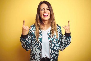 Genç güzel kadın sarı izole arka plan üzerinde rahat ceket giyen elleri ile rock sembolü yapıyor deli ifade ile bağırarak. Müzik yıldızı. Ağır müzik konsepti.