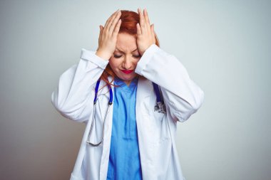 Genç kızıl saçlı doktor kadın beyaz izole arka plan üzerinde stetoskop kullanarak baş ağrısı umutsuz muzdarip ve stresli çünkü ağrı ve migren. Eller baş üstünde.
