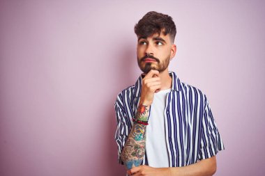 Dövmesi olan genç adam çizgili gömlek giyen çene üzerinde el ile izole pembe arka plan üzerinde duran soru, dalgın ifade düşünme. Düşünceli bir yüzle gülümsüyor. Şüphe kavramı.