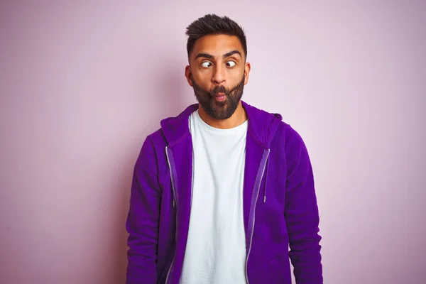 Young indian man wearing purple sweatshirt standing over isolated pink background making fish face with lips, crazy and comical gesture. Funny expression.