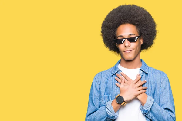 Young african american man with afro hair wearing thug life glasses smiling with hands on chest with closed eyes and grateful gesture on face. Health concept.