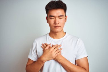 Genç Asyalı Çinli adam kapalı gözleri ve yüzünde minnettar jest ile göğüs üzerinde gülümseyerek izole beyaz arka plan üzerinde duran t-shirt giyiyor. Sağlık kavramı.