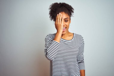 Bir gözünü elle kaplayan izole beyaz arka plan üzerinde duran lacivert çizgili t-shirt giyen Afro-Amerikalı kadın, yüzünde kendine güvenen gülümseme ve sürpriz duygu.