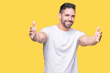 Sarılmak için kollarını açarak gülümseyerek kameraya bakarak izole arka plan üzerinde rahat beyaz t-shirt giyen genç adam. Neşeli ifade kucaklayan mutluluk.