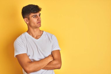 Beyaz tişört giyen genç hintli adam izole sarı arka plan üzerinde duran kolları ile yan bakarak ikna ve kendinden emin geçti