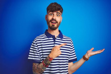 Çizgili polo giyen dövmegiyen genç adam izole mavi arka plan üzerinde duran hayran ve el ile sunarken kameraya gülümseyerek ve parmak ile işaret.