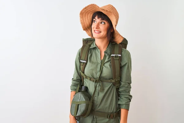 Hiker woman wearing backpack hat and water canteen over isolated white background looking away to side with smile on face, natural expression. Laughing confident.