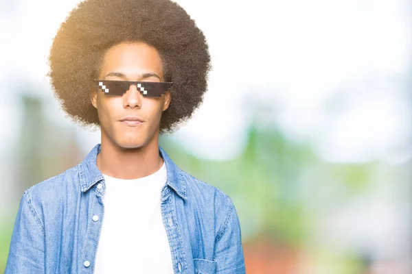 Young african american man with afro hair wearing thug life glasses with serious expression on face. Simple and natural looking at the camera.