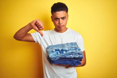 Young brazilian shopkeeper man holding jeans standing over isolated yellow background with angry face, negative sign showing dislike with thumbs down, rejection concept clipart