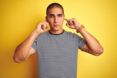 Genç yakışıklı adam yüksek sesle müzik sesi için rahatsız ifade ile parmakları ile kulakları kapsayan sarı izole arka plan üzerinde çizgili t-shirt giyiyor. Sağır kavramı.