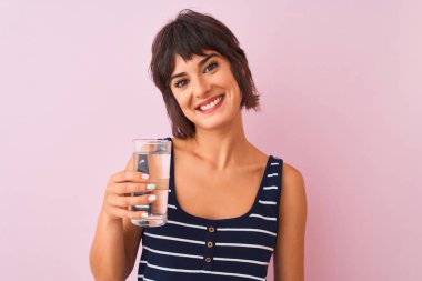 Genç güzel kadın mutlu bir yüz ayakta ve dişleri gösteren kendinden emin bir gülümseme ile gülümseyerek izole pembe arka plan üzerinde duran su cam tutan