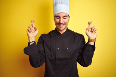 Izole sarı arka plan gesturing parmak üzerinde duran üniforma ve şapka giyen genç şef adam umut ve gözleri kapalı gülümseyerek geçti. Şans ve batıl kavram.