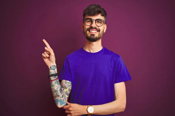 Tシャツとメガネをかけたタトゥーをした若者が 孤立した紫色の背景の上に立ち カメラを見て手と指で指差し 顔に大きな笑みを浮かべている — ストック写真