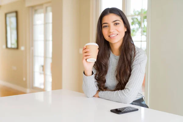 Young woman drinking a cup of coffee at home with a happy face standing and smiling with a confident smile showing teeth