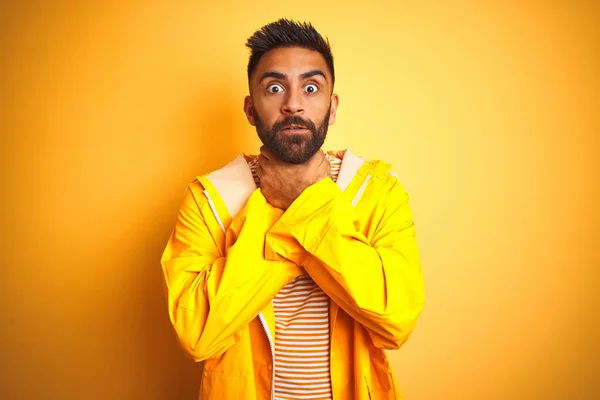 Young indian man wearing raincoat standing over isolated yellow background shouting and suffocate because painful strangle. Health problem. Asphyxiate and suicide concept.