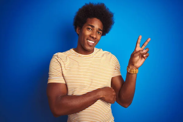 American man with afro hair wearing striped yellow t-shirt over isolated blue background smiling with happy face winking at the camera doing victory sign. Number two.