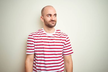 Sakallı genç kel adam beyaz izole arka plan üzerinde rahat çizgili kırmızı t-shirt giyen yan bakarak gülümseyerek ve düşünme uzak bakıyor.