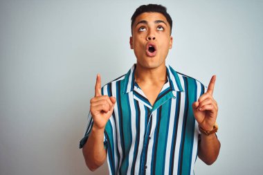 İzole beyaz arka plan üzerinde duran yaz çizgili gömlek giyen genç Brezilyalı adam şaşırttı ve yukarı bakarak ve parmakları ile işaret ve kolları kaldırdı şaşırttı.
