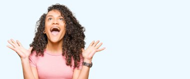 Kıvırcık saçlı Genç güzel kadın pembe t-shirt çılgın kutluyor ve silah yükseltilmiş ve açık gözleri heyecanlı çığlık ile başarı için hayran giyiyor. Kazanan kavramı