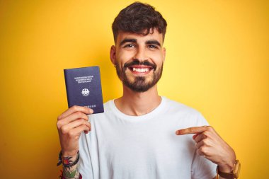 Kendine işaret sürpriz yüzü ile izole sarı arka plan üzerinde Alman Almanya pasaportu giyen dövme li genç adam