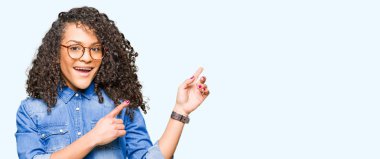 Kıvırcık saçlı Genç güzel bir kadın gözlük gülümseyen ve iki el ve parmak tarafına işaret kamera bakarak giyiyor.