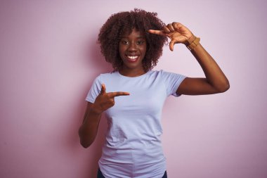 Genç afrikalı afro kadın izole pembe arka plan üzerinde duran t-shirt giyen mutlu yüz ile elleri ve parmakları ile çerçeve yapma gülümseyerek. Yaratıcılık ve fotoğrafçılık konsepti.