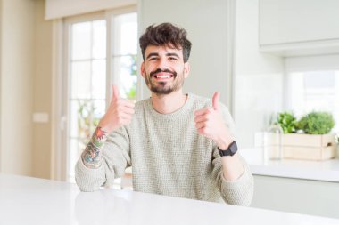 Beyaz masa üzerinde oturan rahat kazak giyen genç adam el ile olumlu jest yapıyor, gülümseyen ve mutlu başparmak. Neşeli ifade ile kameraya bakmak, kazanan jest.