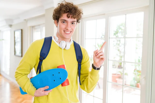 年轻的学生男子戴着耳机和背包拿着滑板很高兴指着手和手指的一边 — 图库照片