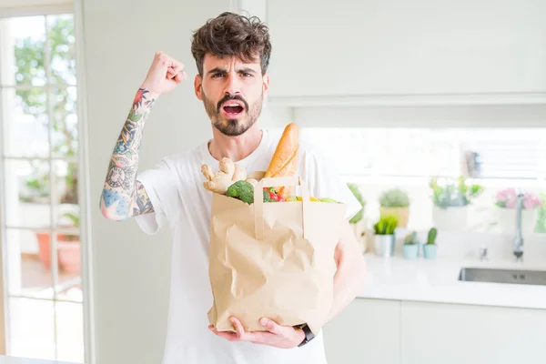 Young man holding paper bag of fresh groceries from the supermarket annoyed and frustrated shouting with anger, crazy and yelling with raised hand, anger concept