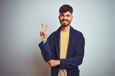 Dövme giyen genç adam izole beyaz arka plan üzerinde duran mutlu yüz zafer işareti yapıyor kameraya göz kırparak gülümseyerek bornoz giyen. İki numara.