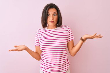 Genç güzel kadın çizgili t-shirt izole pembe arka plan üzerinde duran giyen habersiz ve kollar ve eller kaldırdı ile karışık ifade. Şüphe kavramı.
